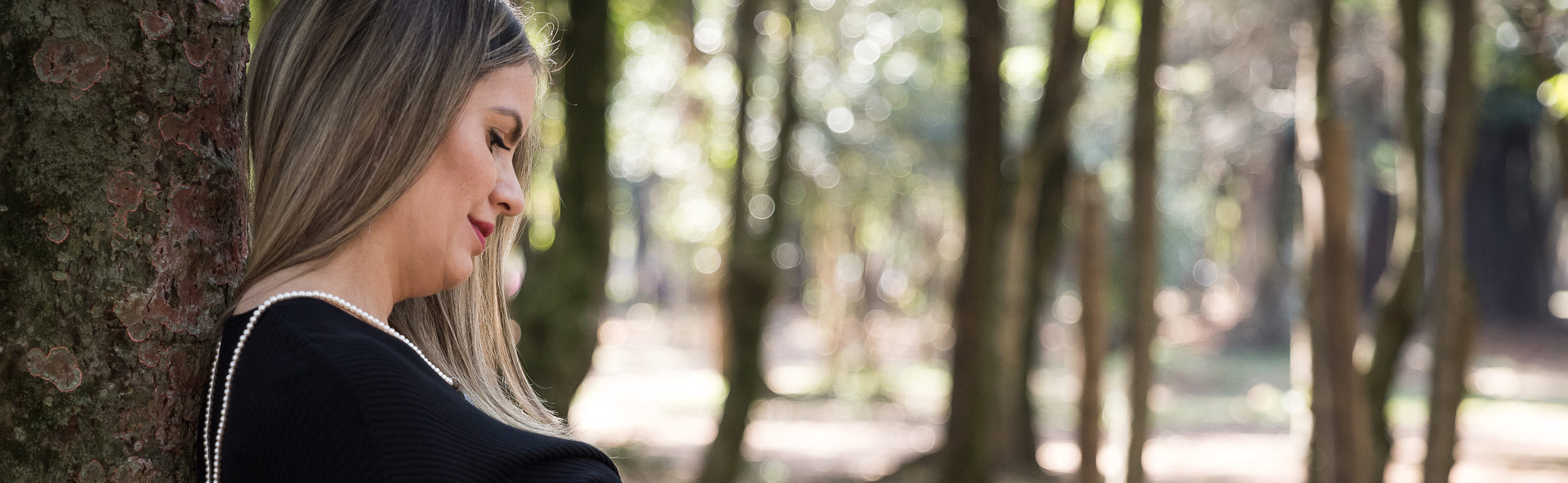 Mulher, grávida encostada em árvore olhando para baixo em direção à barriga