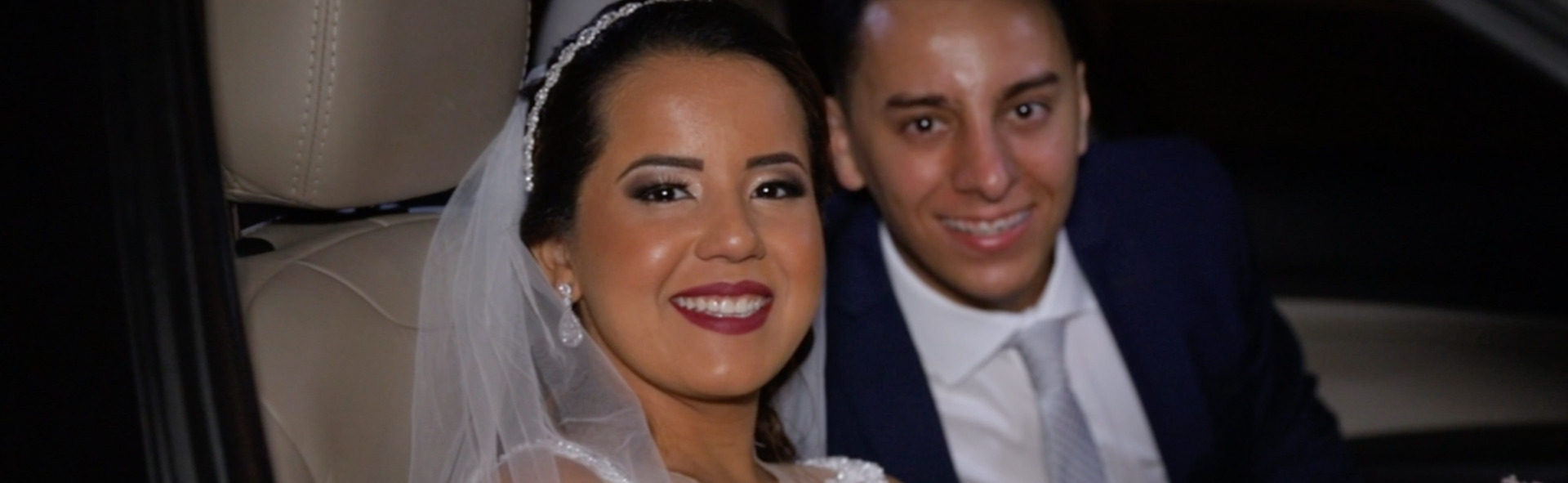 Casal de noivos, recém casados, no carro, saindo para festa. Pode-se vê-los pela janela do passageiro. Ambos sorrindo