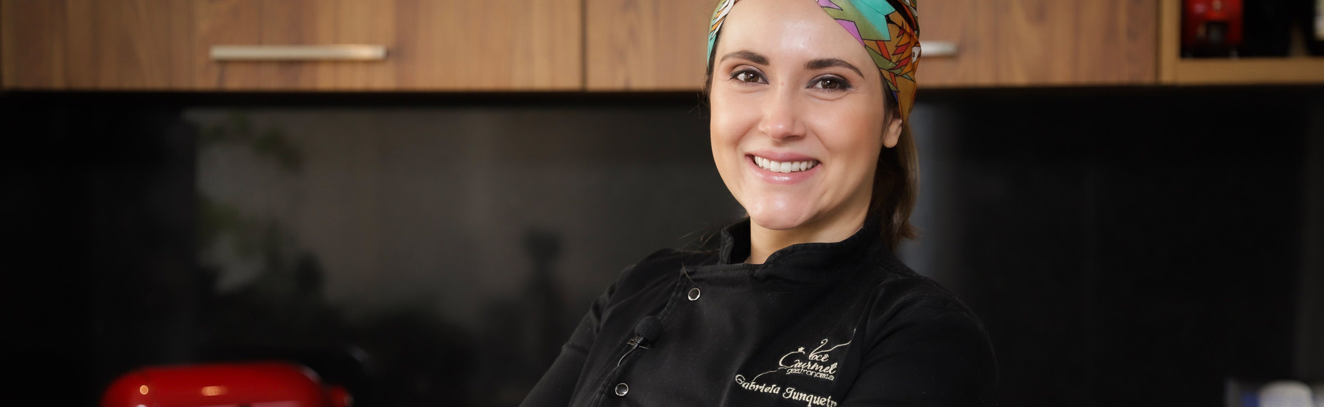 Close da Chef Gabriela, vestida com uniforme preto que leva sua assinatura e também uma faixa de pano colorida na cabeça