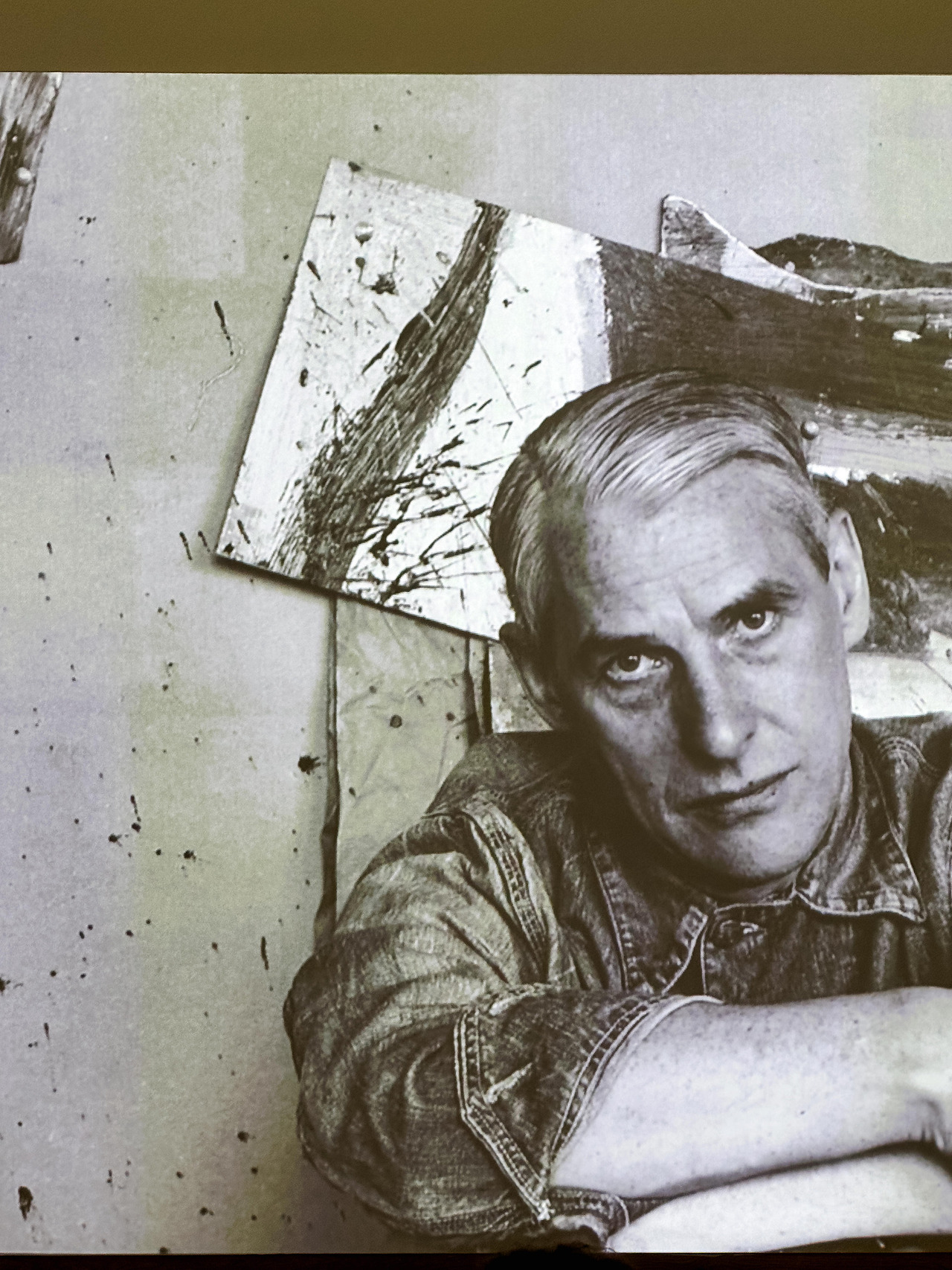 Black-and-white archival portrait of Willem de Kooning sitting in front of one of his paintings.