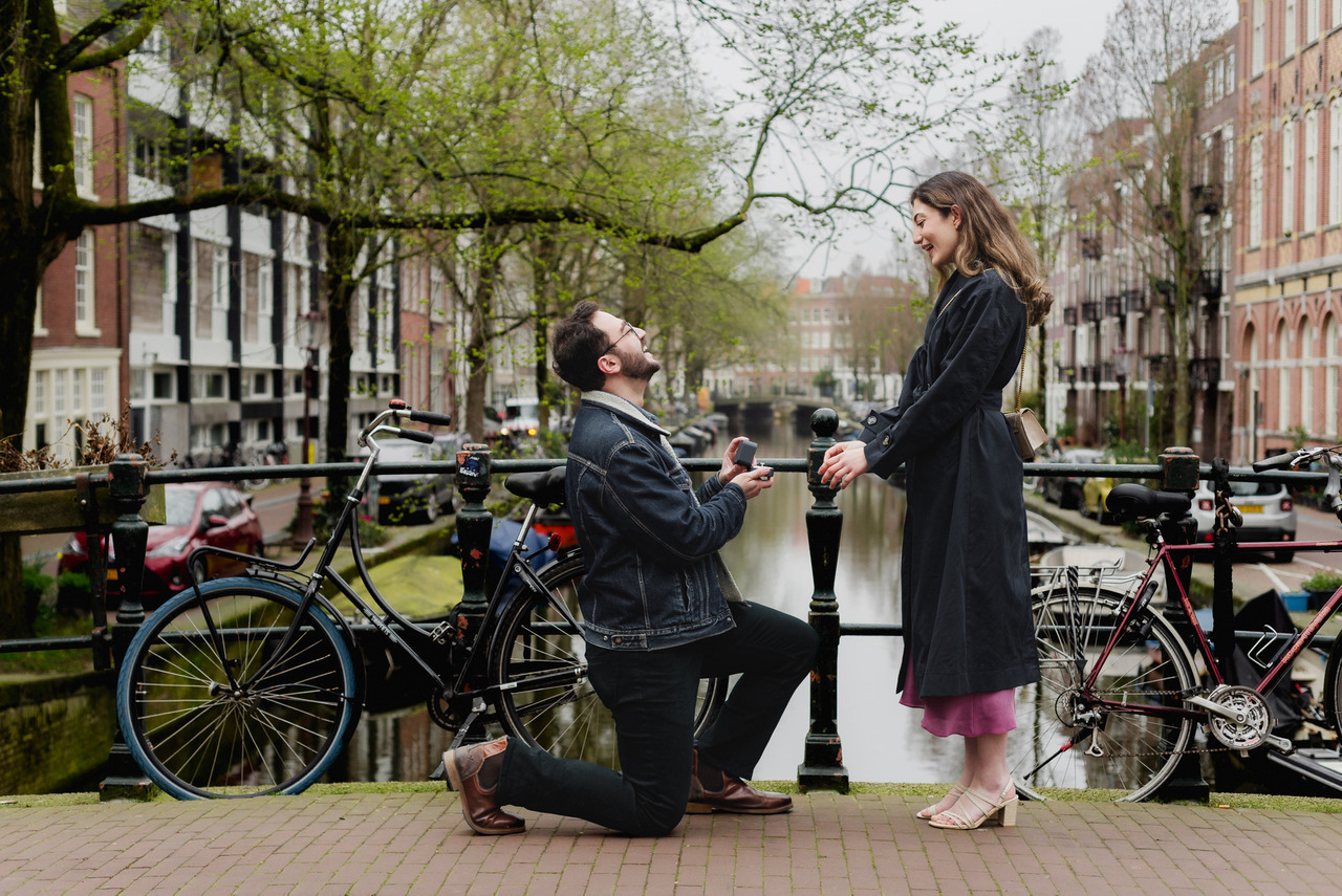 Man kneeling to propose to his partner on an Amsterdam canal bridge, with bicycles and historic houses behind them.