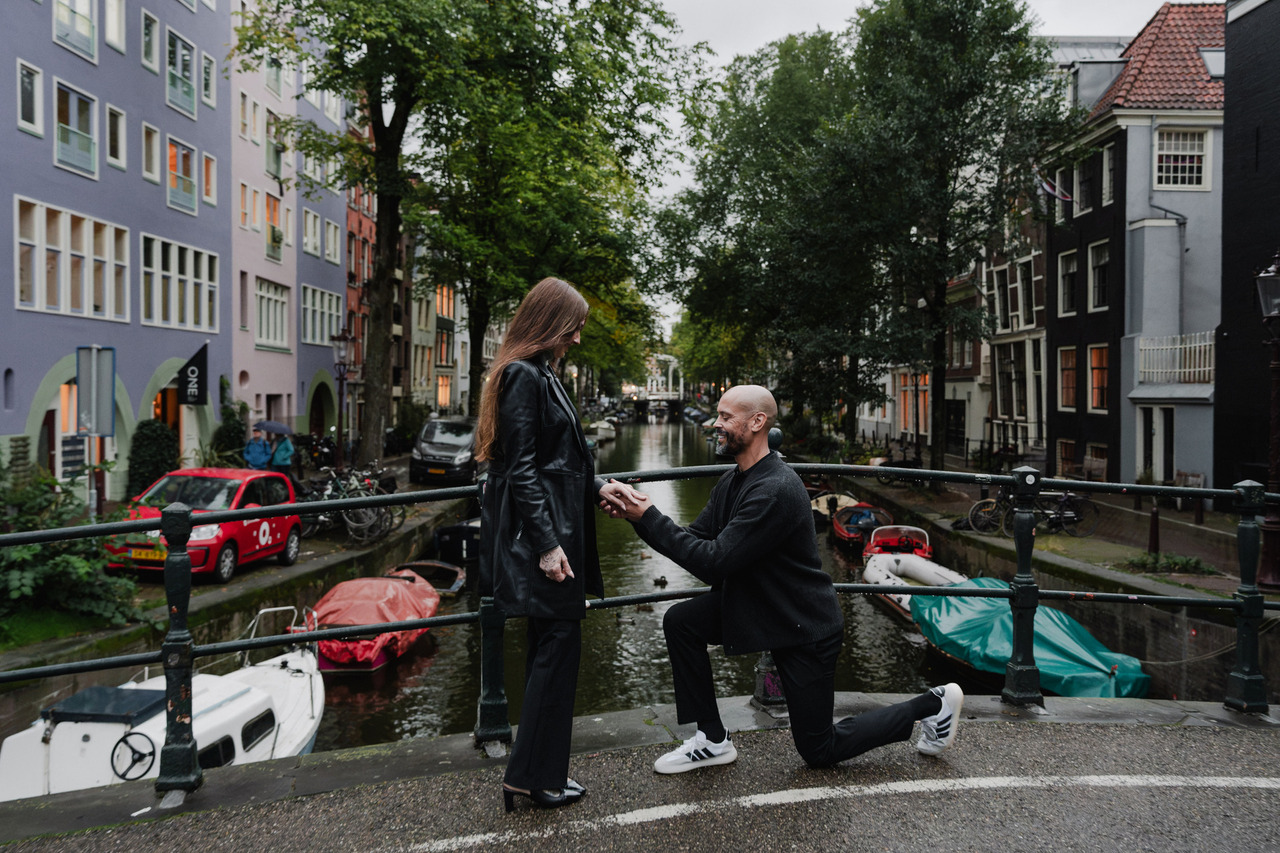 Man proposing to his partner on a canal bridge in Amsterdam, with boats and historic houses behind them.