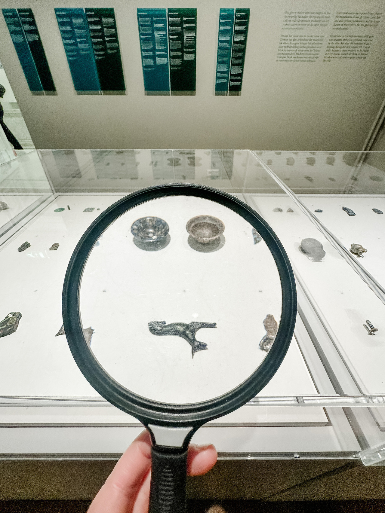 Magnifying glass held in front of small ancient glass fragments displayed in a museum case.