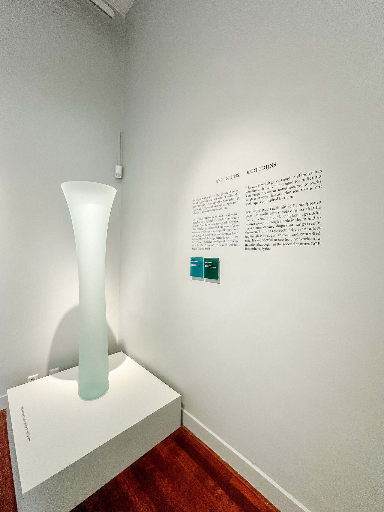 Tall, translucent pale-green glass sculpture by Bert Frijns displayed on a white pedestal at the Allard Pierson Museum.