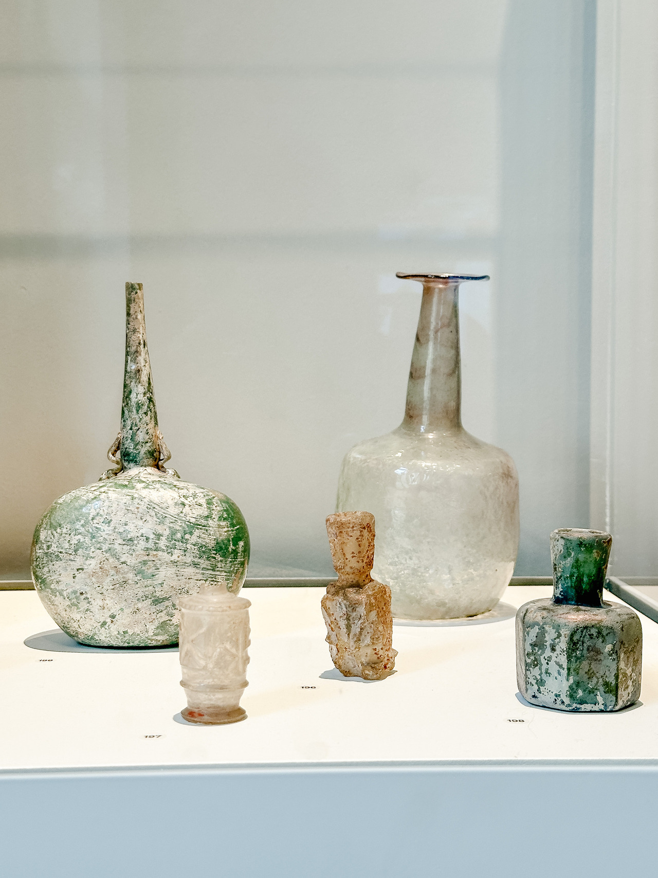 Five ancient glass bottles and small vessels with green, white and iridescent weathered surfaces displayed in a museum case.
