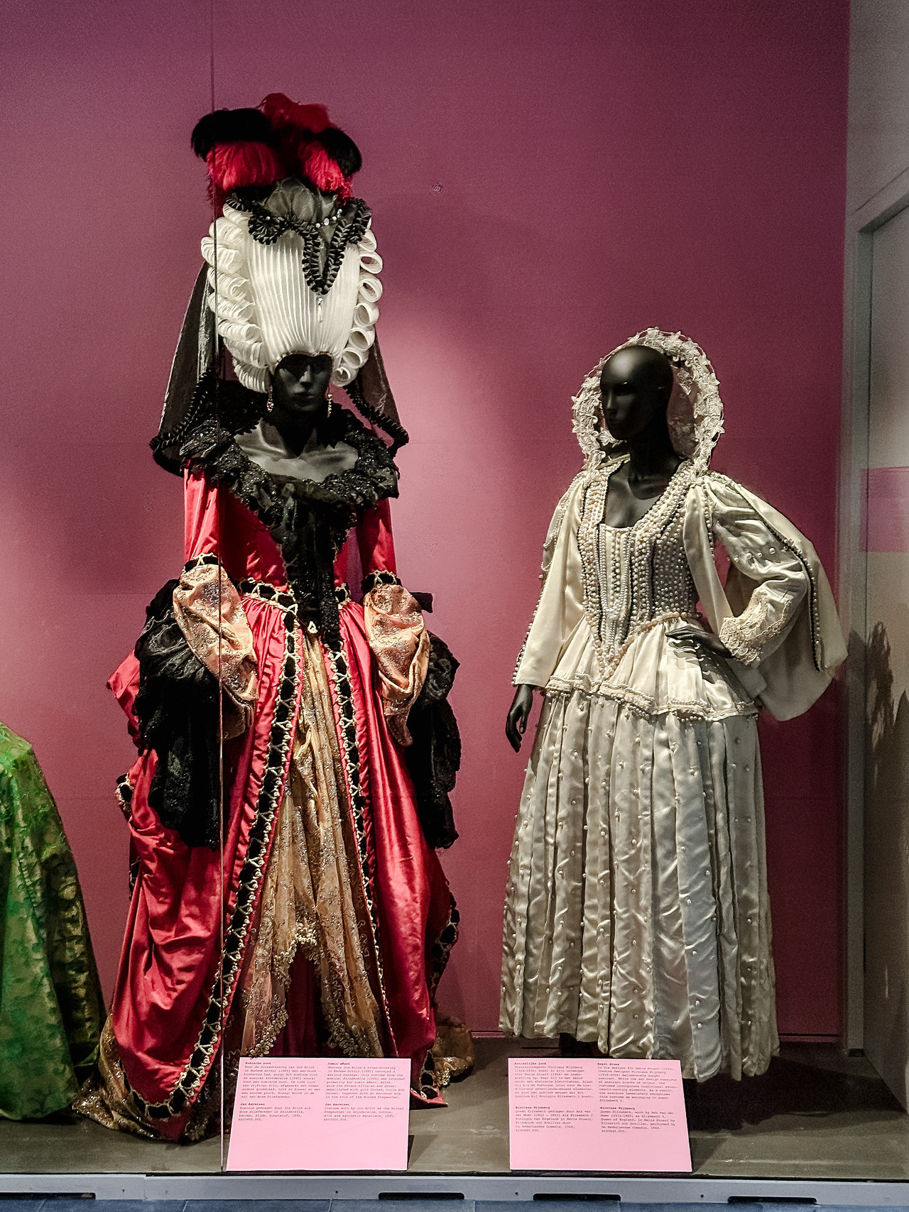Two elaborate historical-style theatrical costumes on mannequins, one red and black with a tall headdress and the other white with pearl-like embellishments.