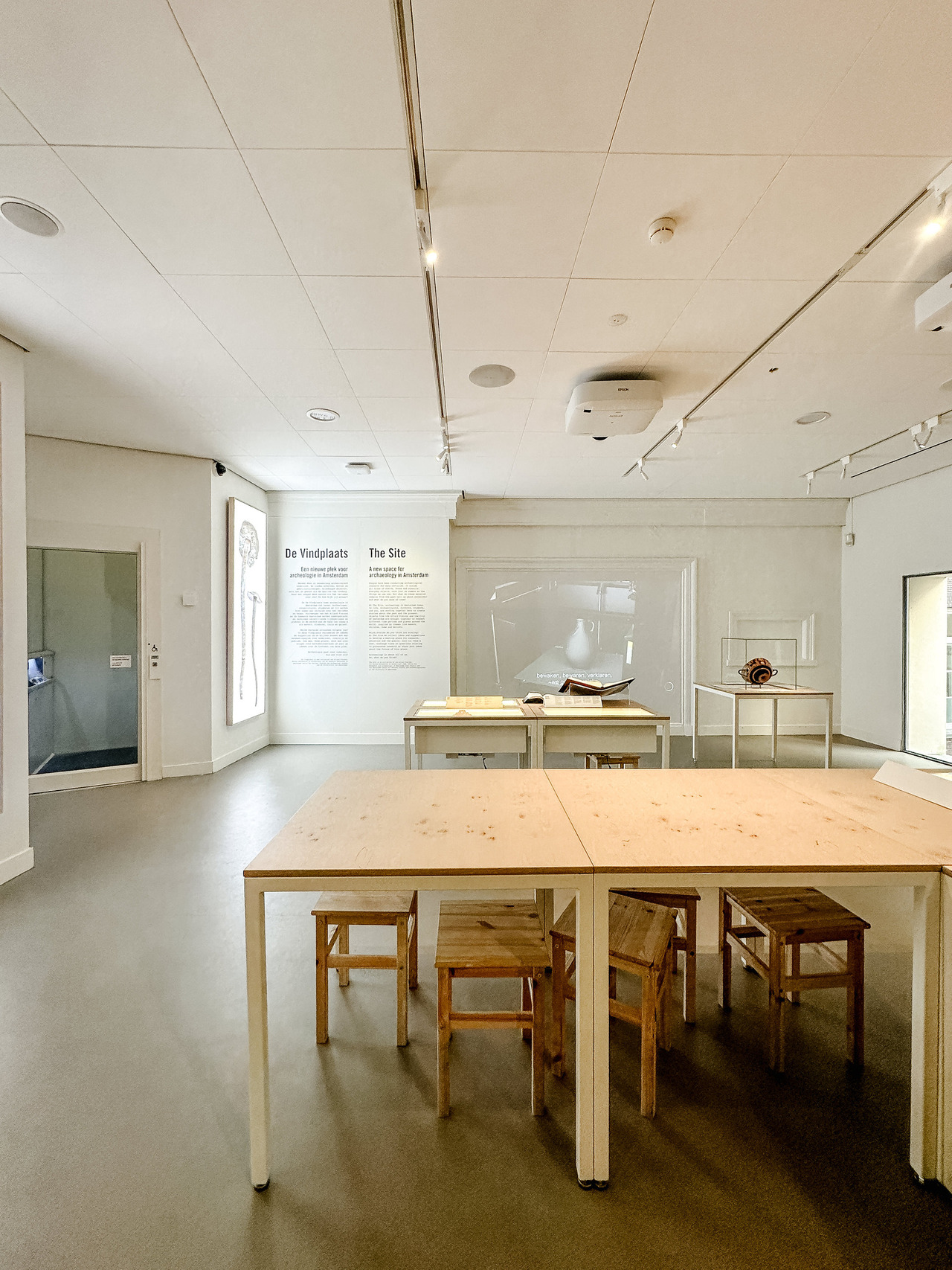 Interior of The Site archaeology exhibition at the Allard Pierson Museum in Amsterdam, with wooden tables, stools and introductory displays.