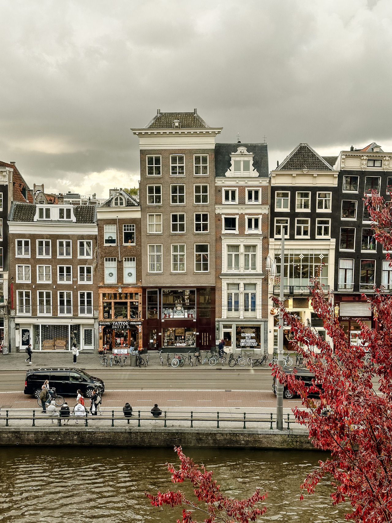 View from the Allard Pierson Museum across an Amsterdam canal towards historic canal houses, bicycles and a red autumn tree beneath a cloudy sky.