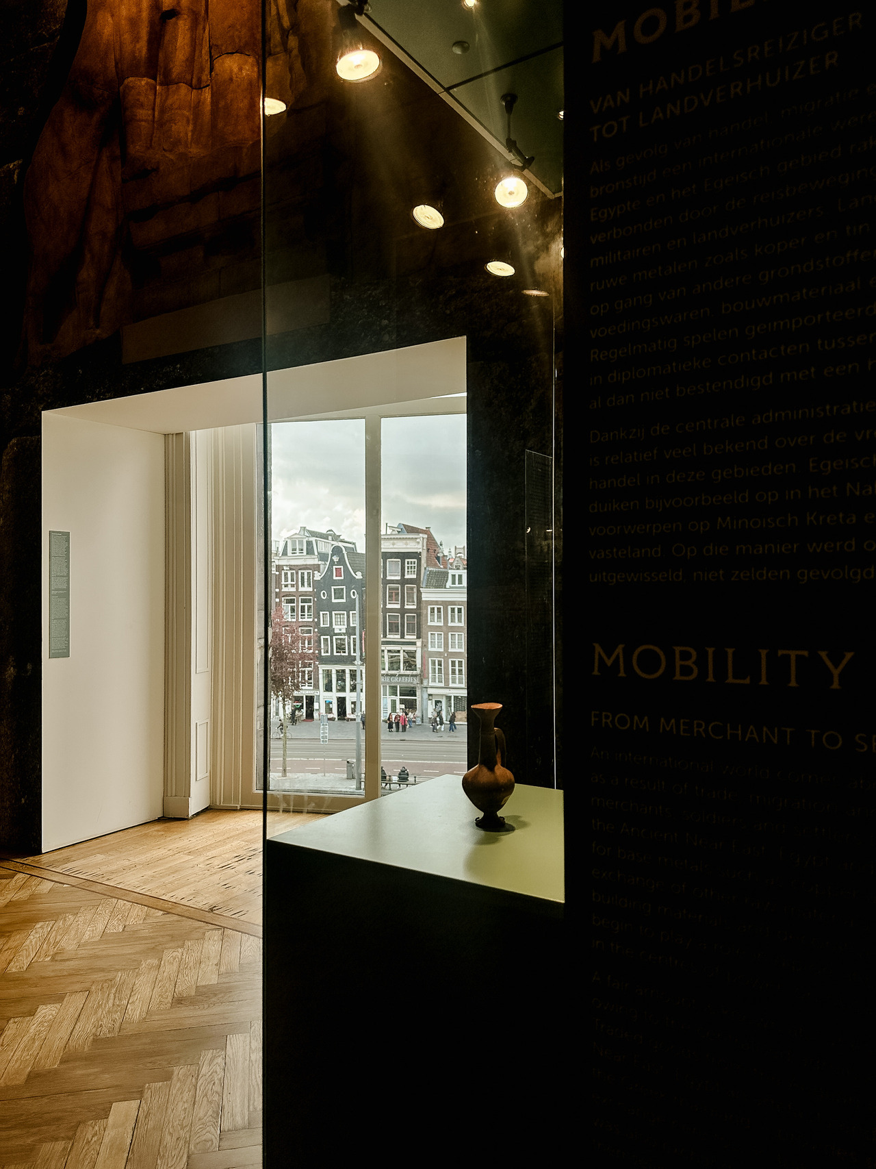 Dark museum gallery with a pottery vessel, a display about ancient trade and a window overlooking Amsterdam canal houses.