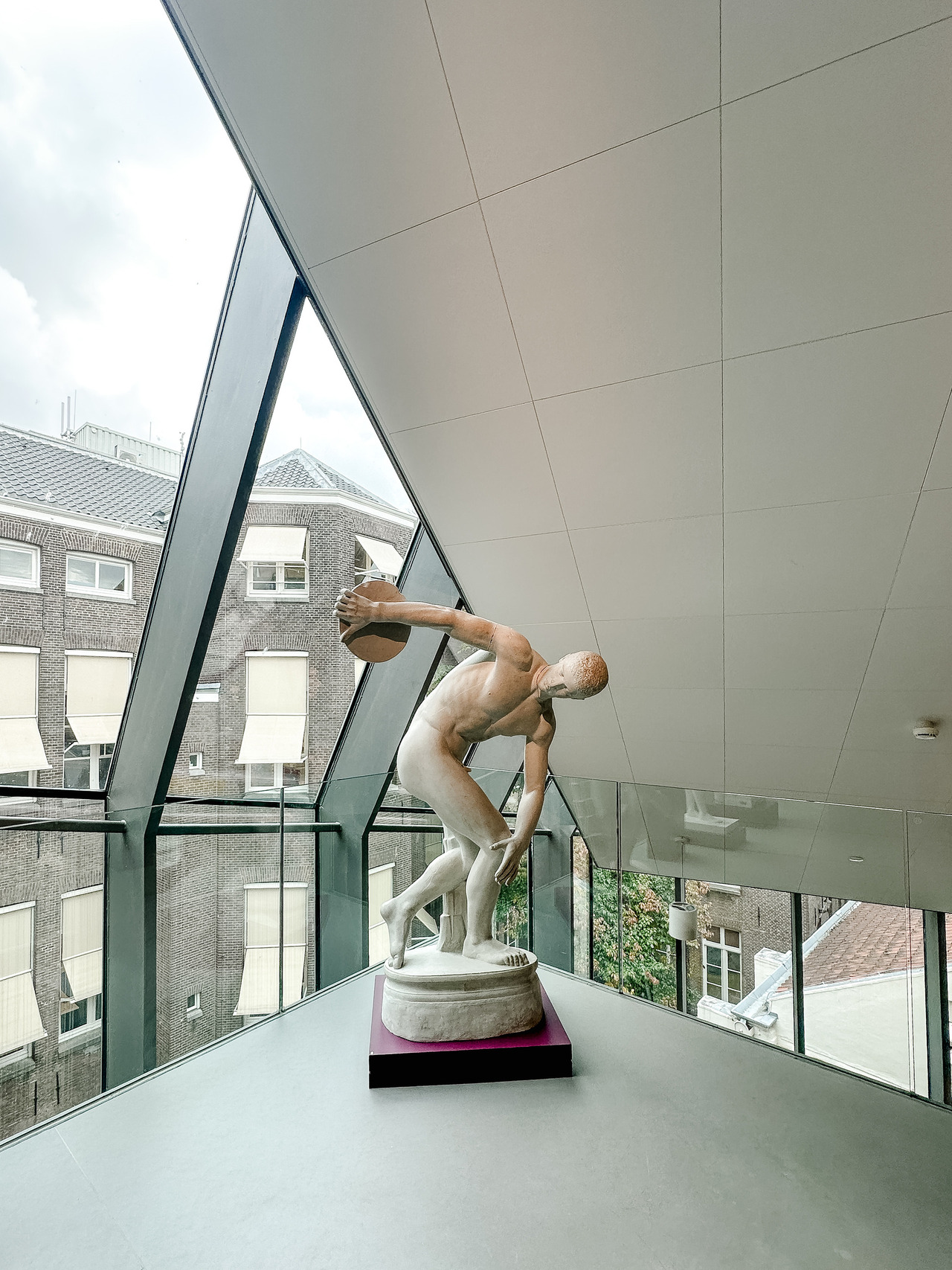 Classical-style sculpture of a discus thrower displayed beside large sloping windows inside the Allard Pierson Museum in Amsterdam.