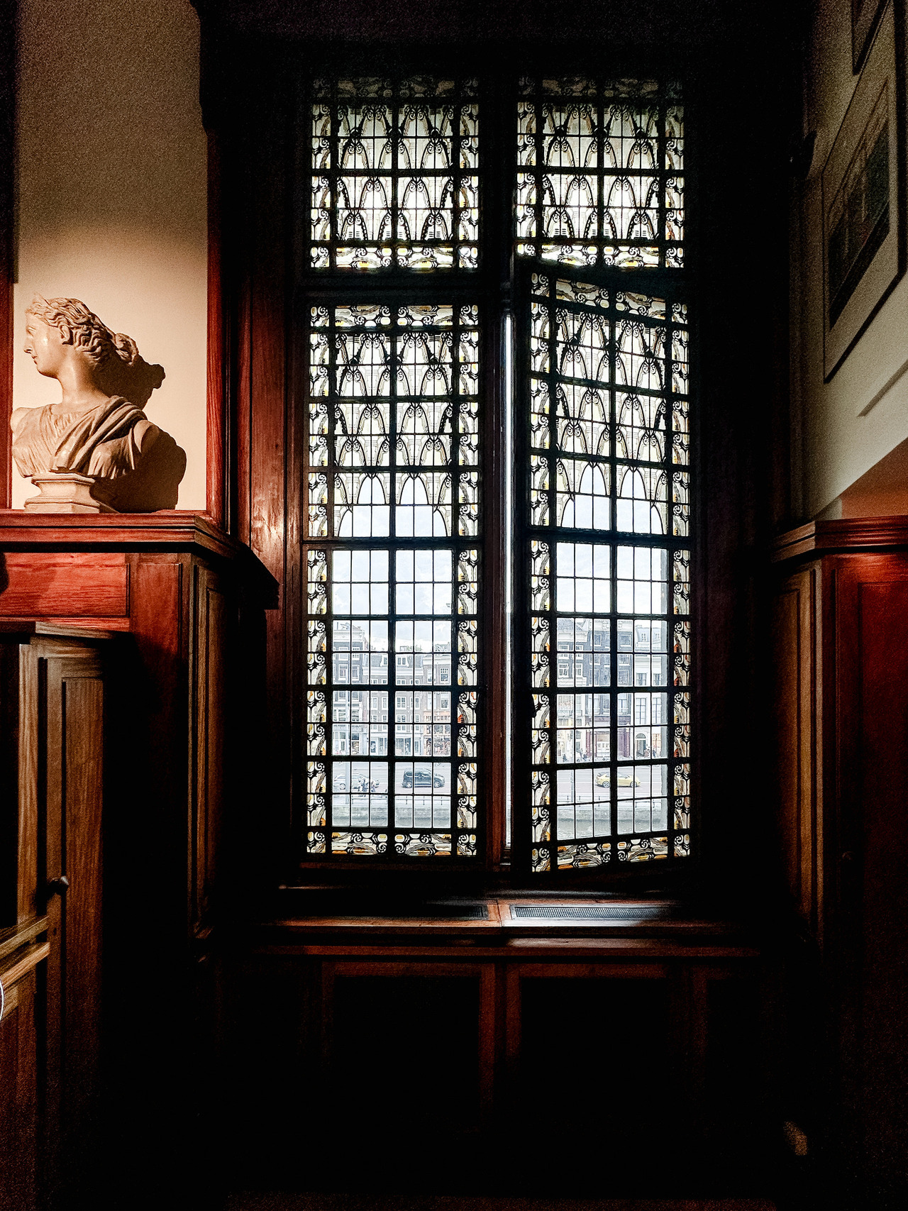 Ornate leaded-glass window framed by dark wooden panelling, with a classical bust on a pedestal inside the Allard Pierson Museum.