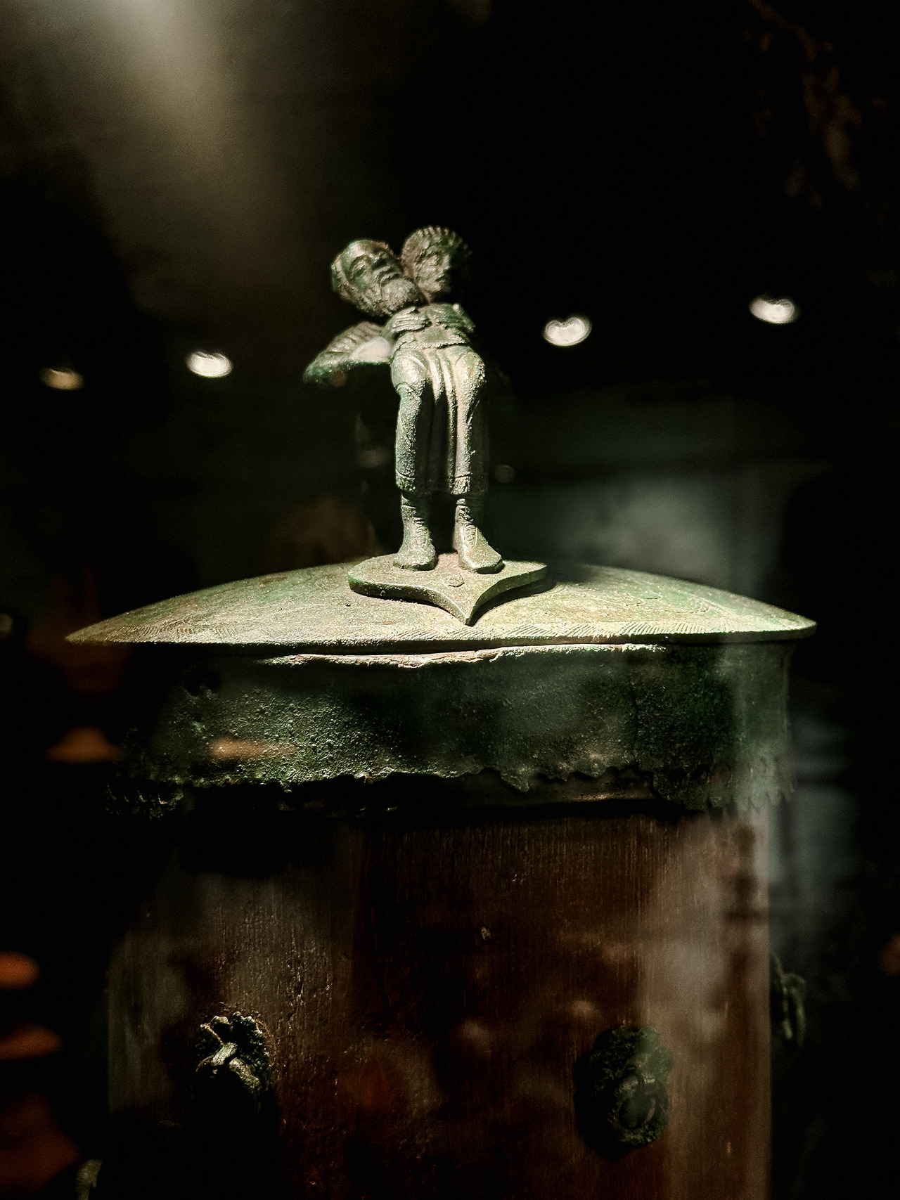 Close-up of a dark bronze vessel with a small human-shaped figurine decorating its lid, displayed in a dimly lit museum case.