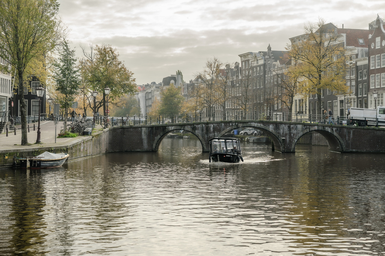 Stone bridge crossing an Amsterdam canal, with a passing boat, historic canal houses and autumn trees beneath a cloudy sky.