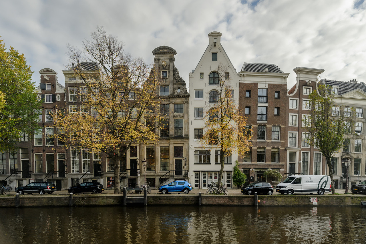 Row of historic Amsterdam canal houses with decorative gables and yellow autumn trees reflected in the canal.