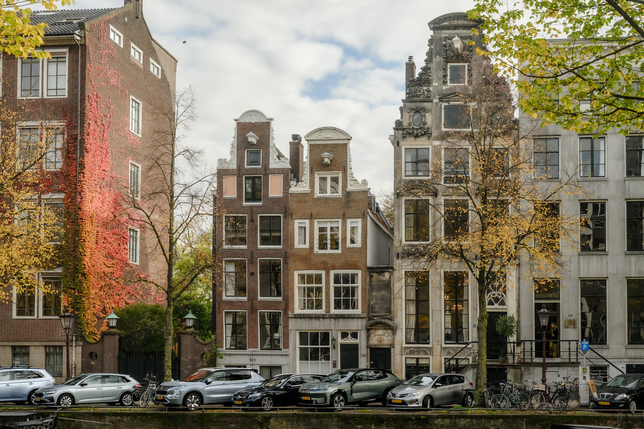 Amsterdam canal houses with ornate gables, a brick wall covered in red autumn foliage and yellow leaves framing the scene.