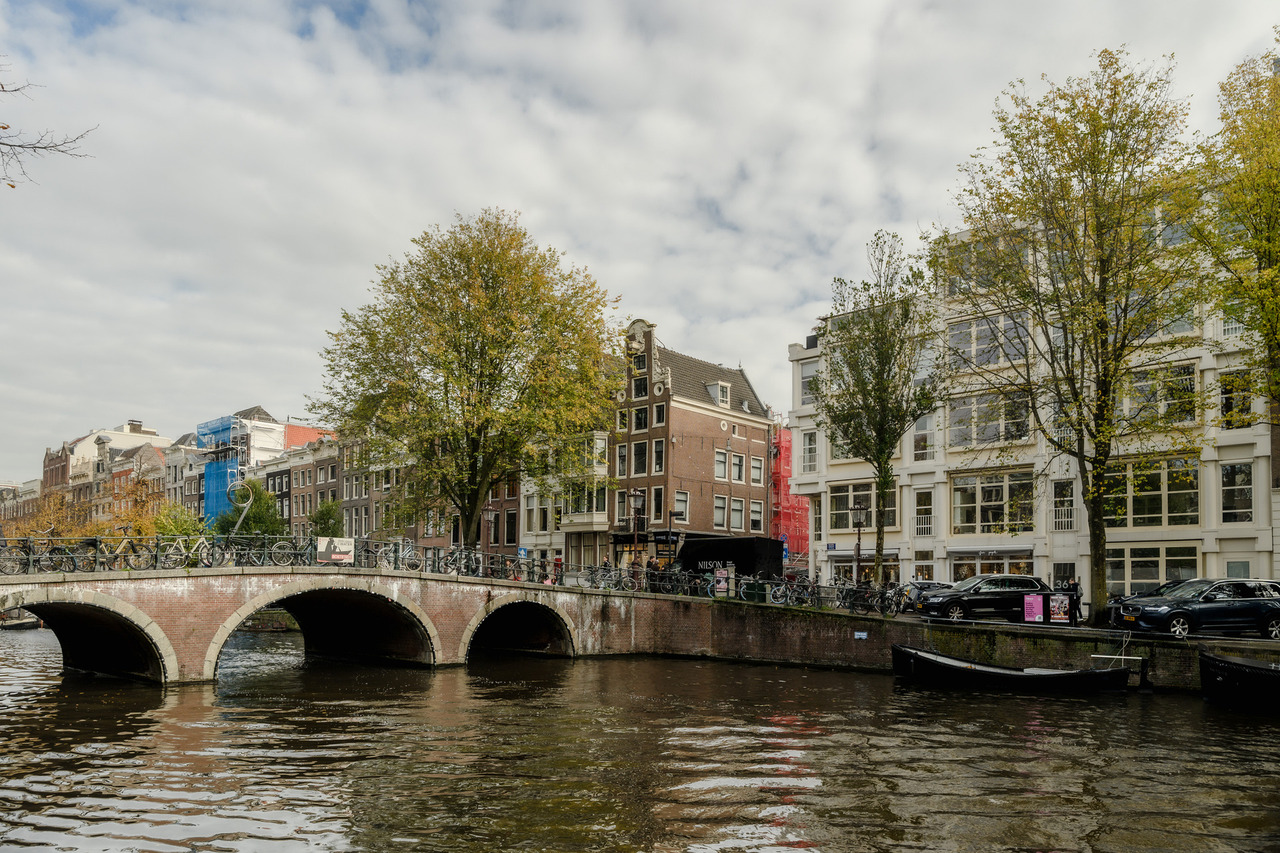 Three-arched brick bridge lined with bicycles crossing an Amsterdam canal beside historic buildings and autumn trees.