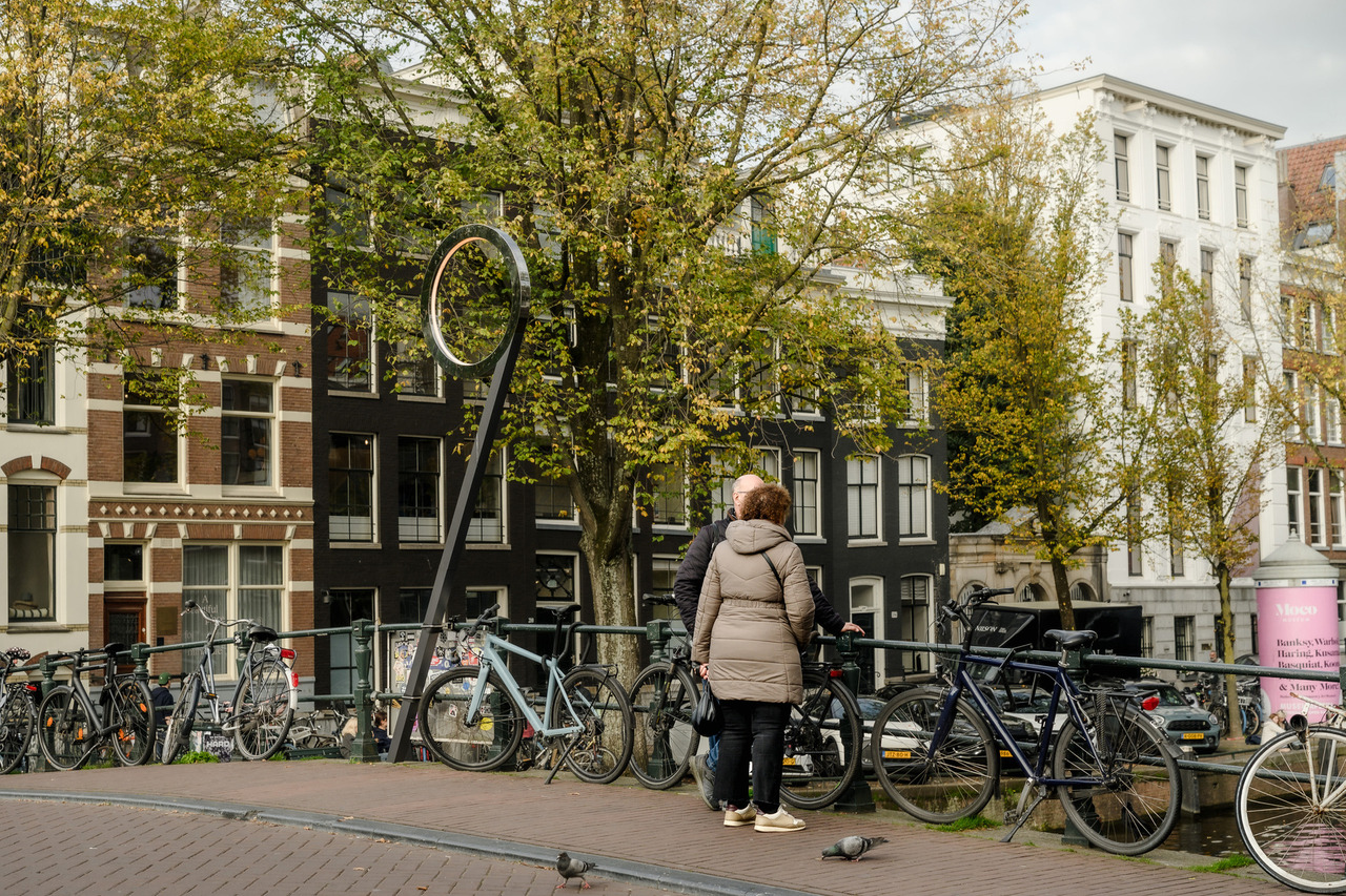 Two people standing beside parked bicycles and a tall circular metal sculpture on an Amsterdam canal bridge in autumn.