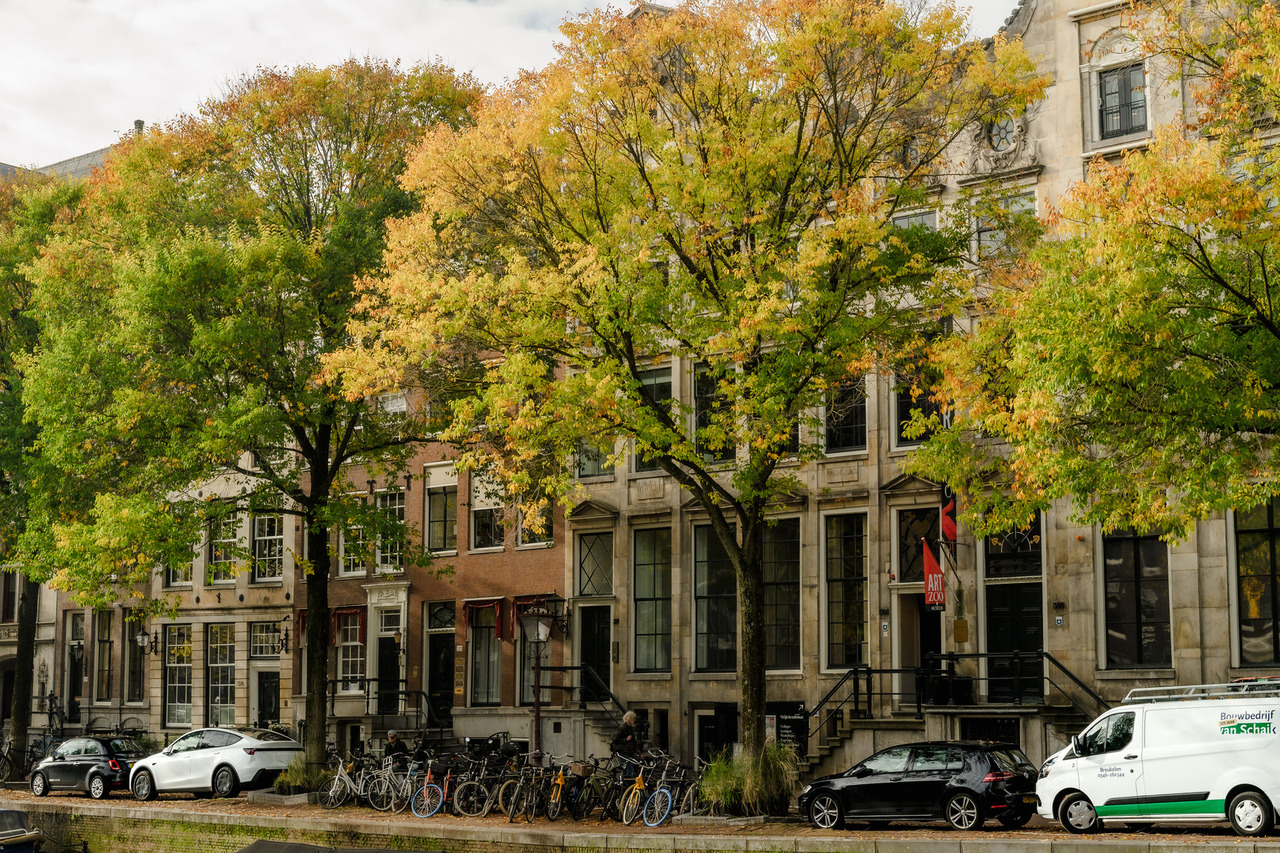 Green, yellow and orange autumn trees lining a row of historic Amsterdam buildings, with bicycles parked beneath them.