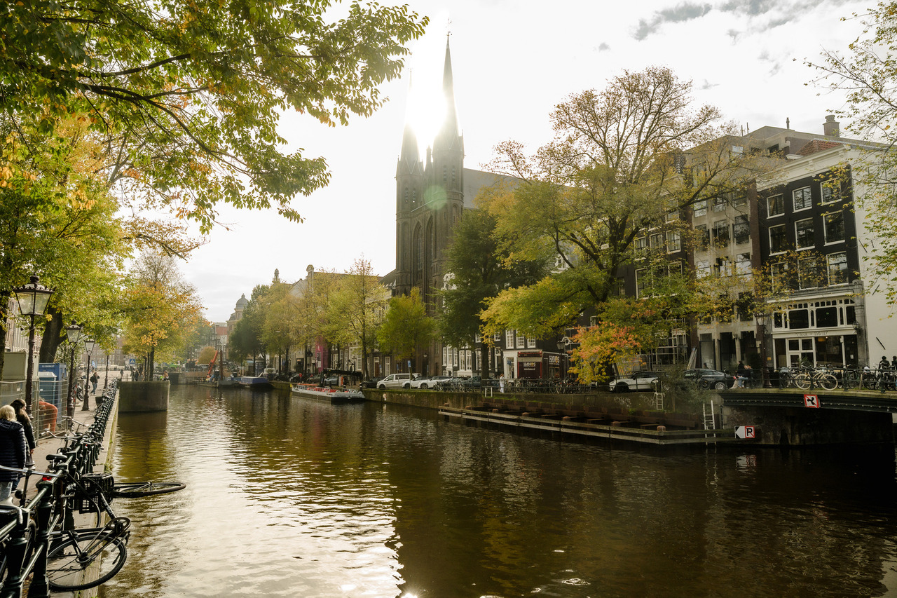 Amsterdam canal with bicycles, autumn trees and a tall church spire silhouetted against bright cloud-filtered sunlight.