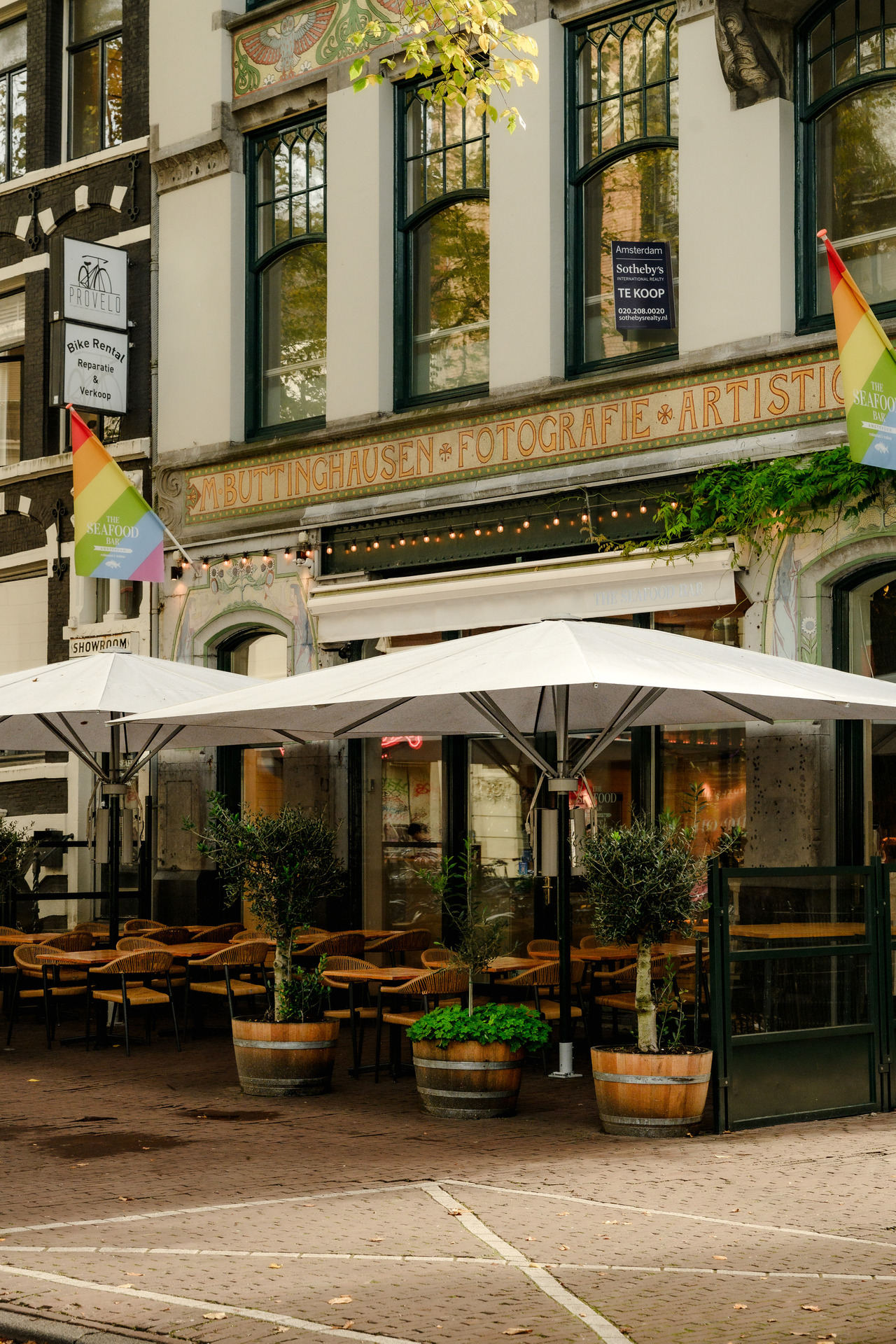 Historic Amsterdam building with decorative lettering reading Buttinghausen Fotografie Artistiek above a café terrace with white umbrellas.