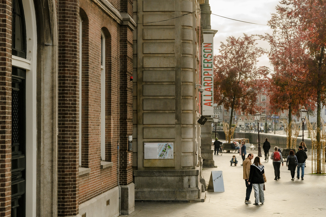 Side entrance approach to the Allard Pierson Museum in Amsterdam, with its vertical museum sign, stone façade, pedestrians and red autumn trees.