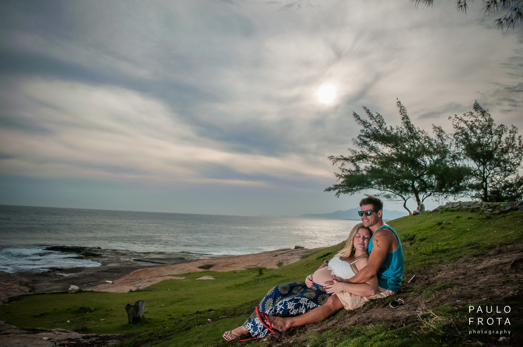 casal gestante em ensaio em saquarema se abraça sentados na grama olhando para o mar.