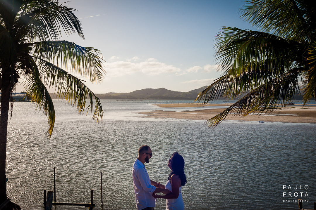 casal de mãos dadas, riem. ao fundo a lagoa de saquarema e os coqueiros nas duas laterais da imagem.