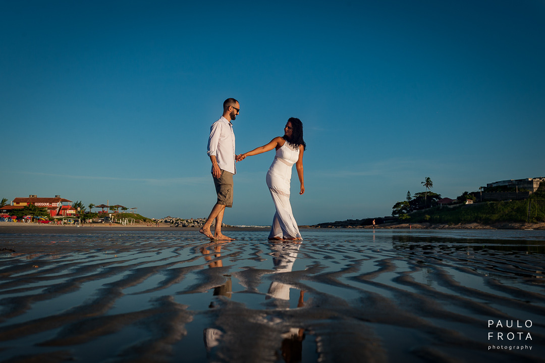 casal passeando pela areia molhada da lagoa de saquarema. ela vai na frente de mão dada com seu par, puxando ele.