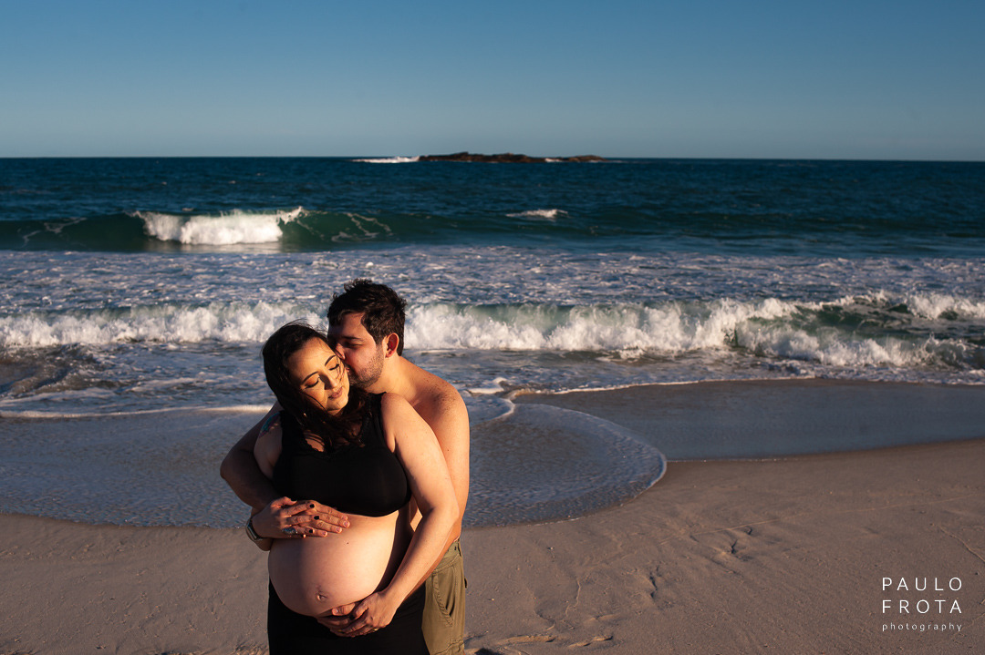 ele abraça ela enquanto o sol bate nos dois pela lateral direita. ela de olhos fechados