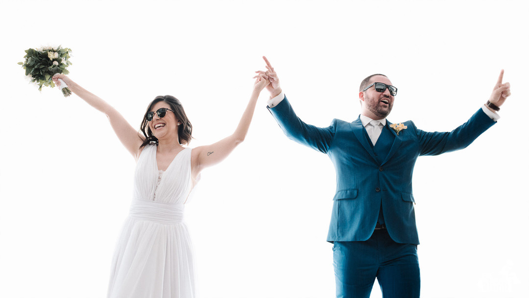 fotografia de casal comemorando, de braços erguidos, felizes, sorrindo, de óculos escuros, num fundo branco