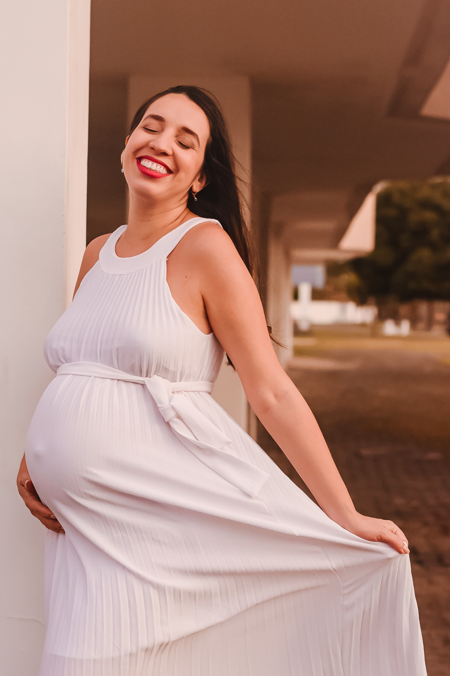 gestante posando com vestindo branco para ensaio de gestante em local externo. ela está´com os olhos fechados e sorri