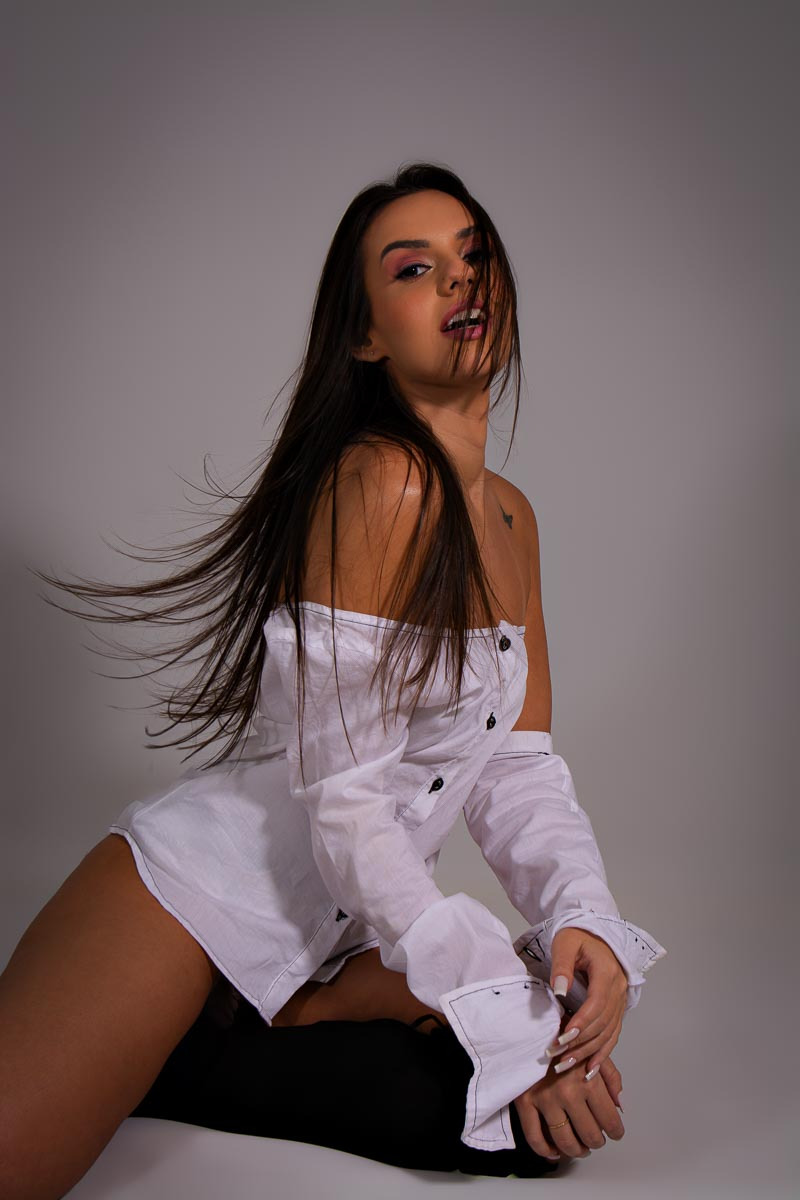 ensaio feminino sensual em estúdio mulher sentada no chão do estúdio olha para a camera vestindo blusa social branca e os cabelos tem movimento