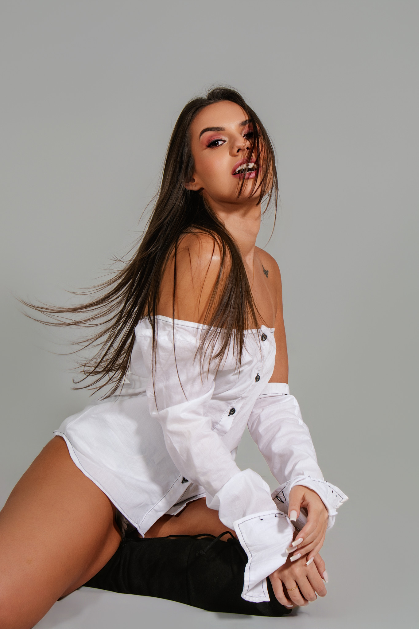 ensaio feminino sensual em estúdio mulher sentada no chão do estúdio olha para a camera vestindo blusa social branca e os cabelos tem movimento