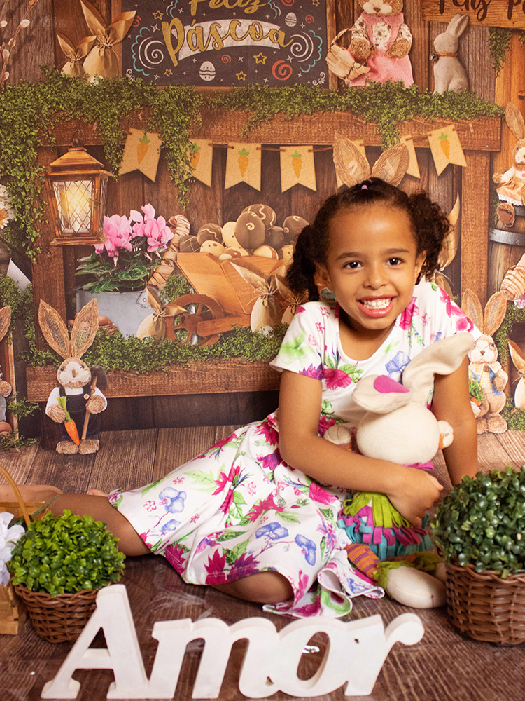 Uma doce menina de cachinhos e cabelos em maria chiquinha está sorrindo segurando um coelho colorido de pelúcia, usa um vestido branco e o fundo é de Páscoa.