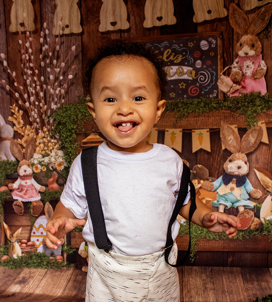  Um lindo bebê pardo de cachinhos castanhos escuros mostrando a língua com dois dentinhos de fora e um sorriso no olhar, ele veste uma camiseta branca, uma bermuda branca listrada e usa suspensórios, o fundo fotográfico é de Páscoa.