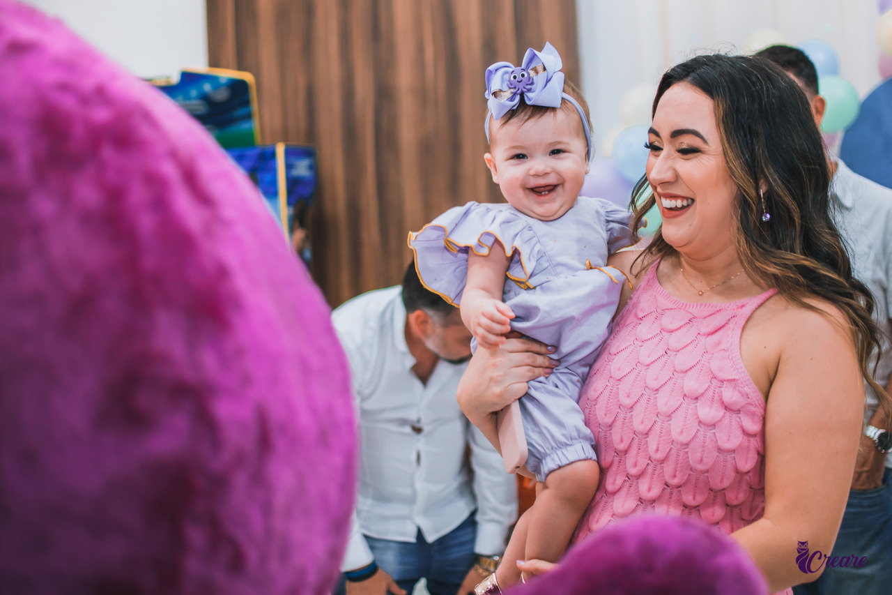 a fotos contem uma criança no colo de sua mãe, as duas estão sorrindo. A criança está com uma roupa lilás bem clarinha e a mãe de roupa rosa, bem clara. 