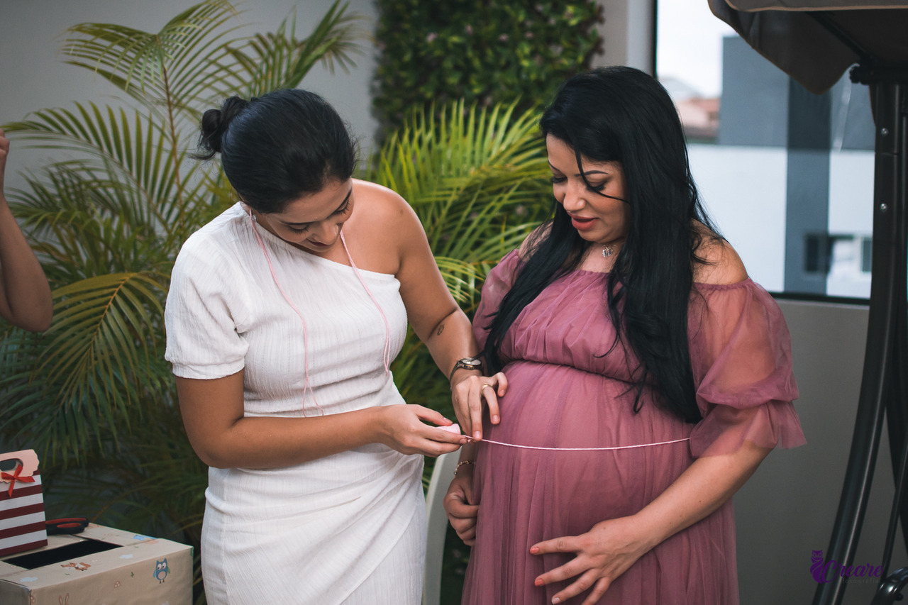 A imagem contém duas mulheres adultas, ambas brancas com cabelos pretos; uma está com um vestido branco e a outra com um vestido rosa.