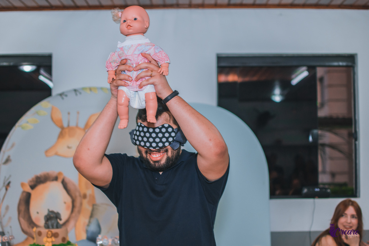 A imagem contém um homem adulto com uma boneca erguida acima da cabeça. O homem veste uma blusa azul e uma venda nos olhos.