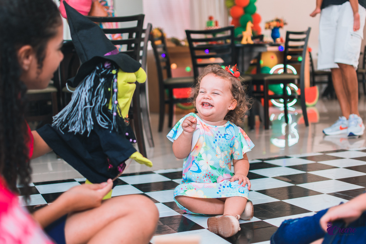 A imagem mostra uma criança pequena sentada no chão de um espaço interno com piso xadrez preto e branco. Ela está usando um vestido colorido com estampas de flores e parece muito feliz, rindo e olhando para alguém que está brincando com ela. Perto da criança, uma pessoa segura um fantoche de mão em formato de bruxa, com um chapéu preto pontudo, cabelo de fios cinza e rosto verde. Ao fundo, há várias cadeiras pretas e decoração de festa, incluindo balões coloridos em verde e laranja. A atmosfera é alegre e divertida, típica de uma festa infantil, com a criança claramente se divertindo com a interação com o fantoche.