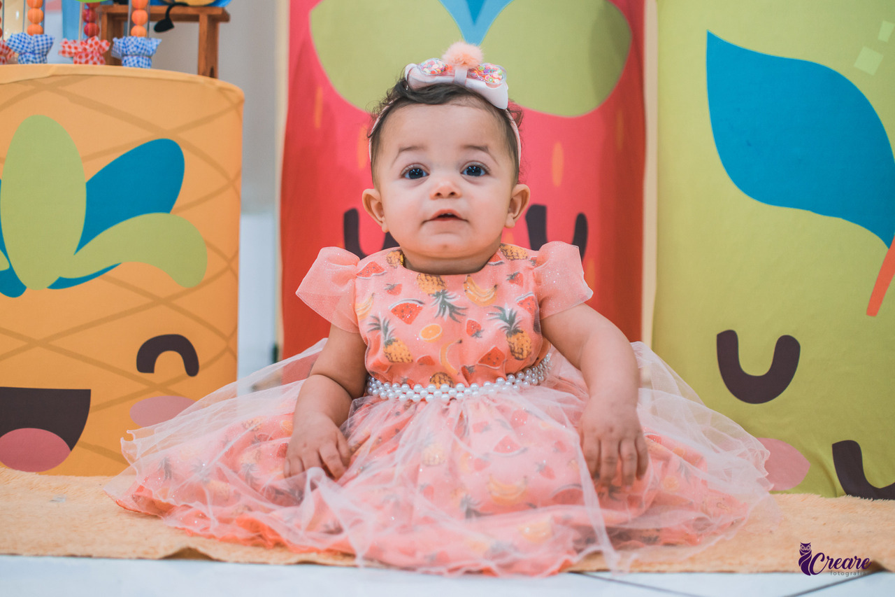 Um bebê está sentado em um tapete bege, com um cenário colorido ao fundo que parece ser de uma festa infantil. Ela usa um vestido rosa com estampas de abacaxi e uma faixa de cabelo combinada. O fundo tem elementos decorativos grandes e coloridos, com desenhos de frutas estilizadas, como um abacaxi. O bebê olha diretamente para a câmera com uma expressão calma.