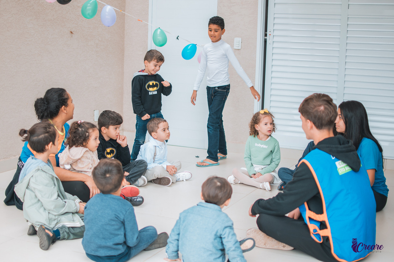 Um grupo de crianças está reunido em um espaço fechado, possivelmente uma varanda ou uma sala com piso branco e paredes bege. Eles estão sentados no chão em um círculo, parecendo participar de uma atividade. Há balões coloridos pendurados acima deles e duas pessoas vestidas com uniformes azuis que parecem ser os organizadores da atividade. As crianças estão atentas, e algumas estão sorrindo.