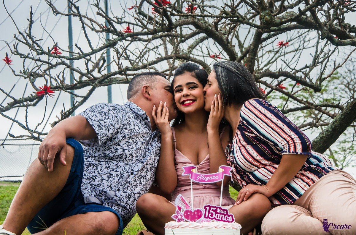 Uma jovem de 15 anos está sorrindo e sentada na grama. Ela está no meio de um homem e uma mulher que estão beijando suas bochechas. Atrás deles, há uma árvore com alguns galhos secos e algumas flores vermelhas. A jovem está segurando um topper de bolo que diz "Nayara, 15 anos" em rosa. Há um bolo de aniversário decorado na frente deles.