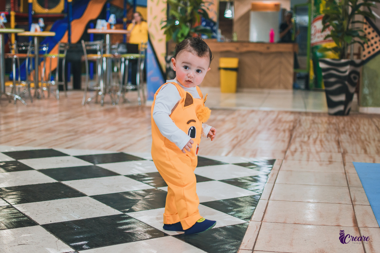 Um bebê usando um macacão amarelo, durante sua festa de aniversário de 1 ano.