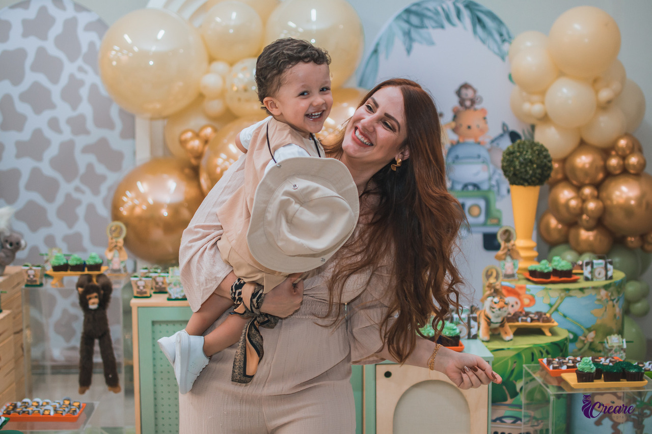 a imagem contém uma mulher segurando um menino de dois anos no colo, os dois sorriem. O menio veste roupas claras e um chapéu, a mulher veste roupas claras e tem cabelos longos.