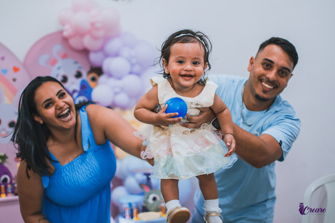 A imagem contém, uma menina de 1 ano de idade, brincando com os pais. Os pais estão sorridentes e vestidos de azul e a criança veste um vestido branco e segura uma bolinha azul nas mãos.