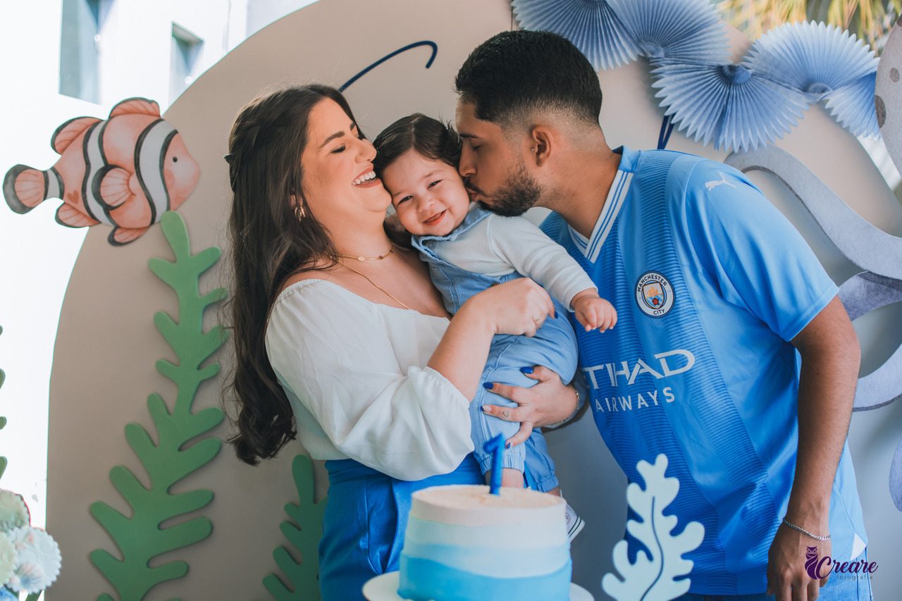 a Imagem contém uma mulher vestida de blisa branca e calça azul segurando um bebê de um ano no colo, e o pai do bebê vestido com uma camisa azul beijando a bochecha do bebê. 