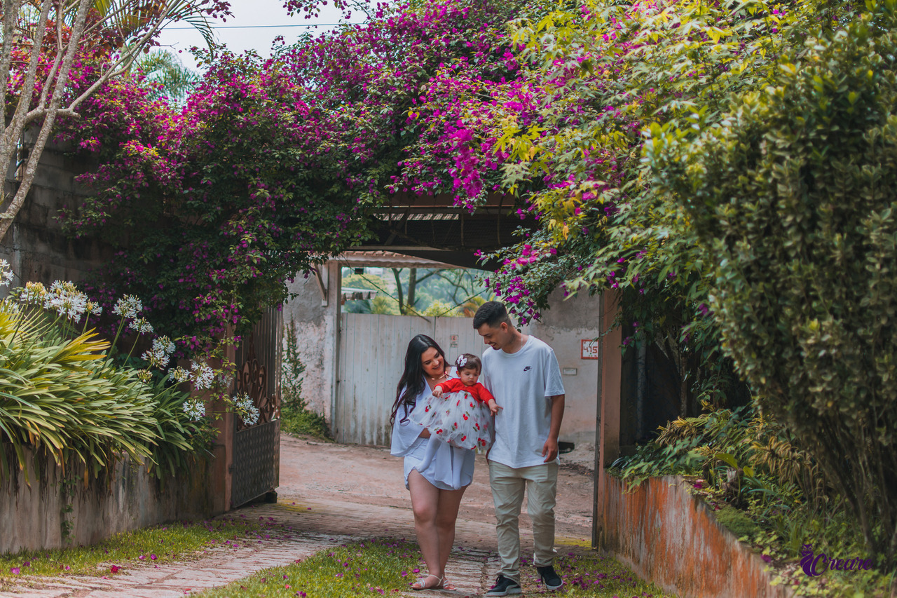 A imagem contém uma mãe e um pai caminhando com sua filha de um ano de idade nos braços. Muito verdade e flores coloridas ao redor.