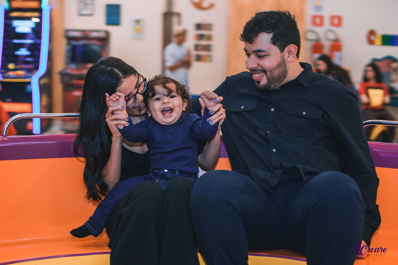 A imagem contém, dois adultos e uma criança sentados em um brinquedo de girar. A criança sorri divertidamente e os adultos olham para o bebê com cara de felicidade