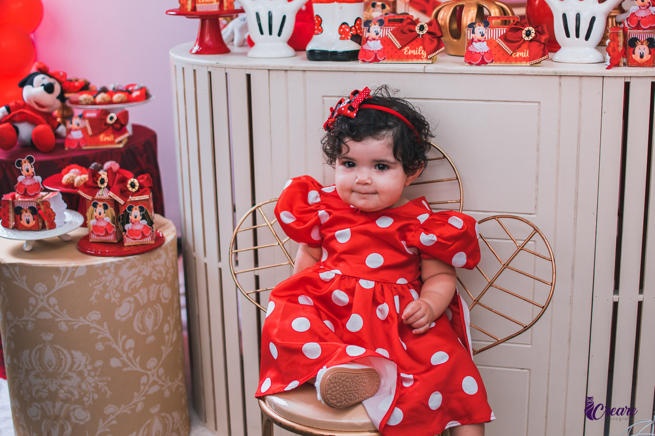 A imagem contém uma menina de um ano de idade, sentada em um cadeira em frente a uma mesa decorada, usando um vestido vermelho e laço vermelho na cabeça. O tema é Minnie.