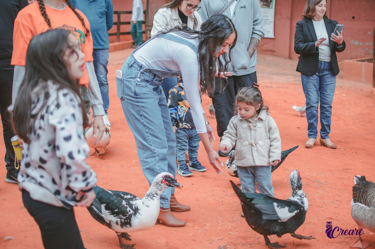 a imagem contém uma criança e uma mulher alimentando alguns patos, a criança está de blusa de frio branca e calça jeans, a mulher veste uma macacão jeans e blusa branca. Em primeiro planos uma criança com blusa branca e calça preta.